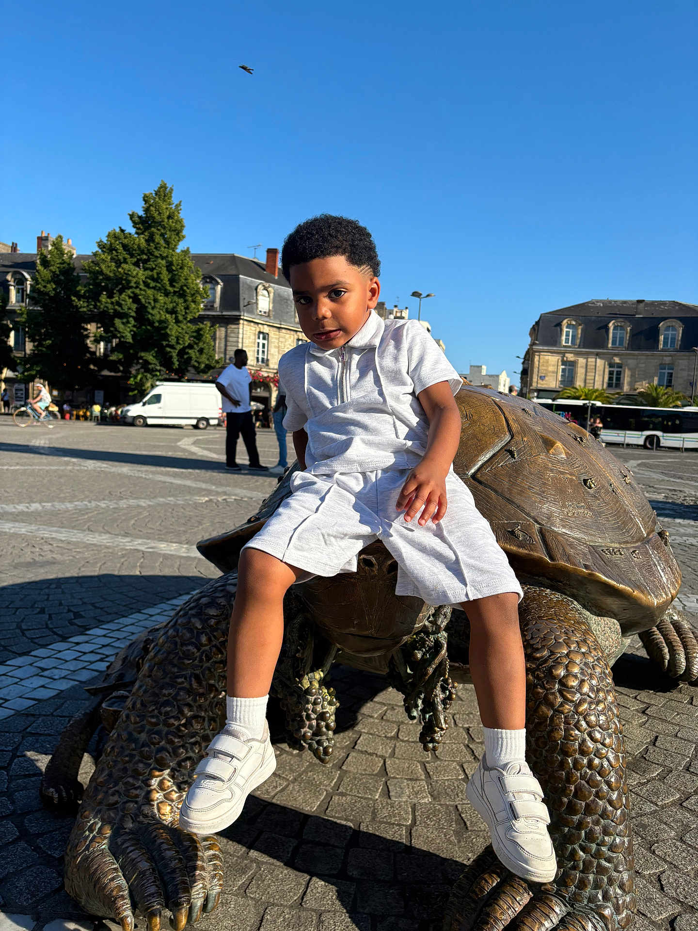 Darian participe au concours pour gagner de l'argent avec cette photo : child, boy, sculpture, turtle, bronze, outdoor, daytime, urban, square, building, sidewalk, people, casual, white_clothing, sneakers, shorts, trees, sky, statue, public_space