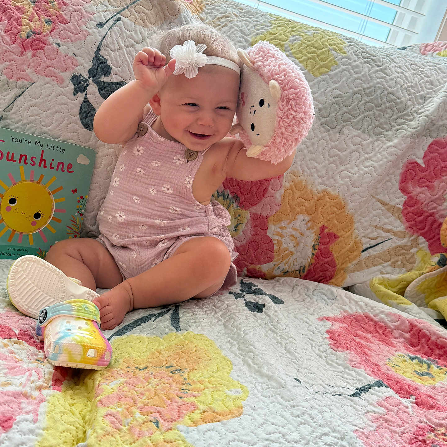 Paisley joined the competition — help win amazing prizes! baby, child, smiling, plush_toy, headband, pink_clothing, quilted_couch, colorful, floral_pattern, children_book, shoe, indoor, window, blinds, happy, sitting, playful, cute, portrait, toddler