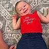 baby, child, smiling, red_top, blue_shorts, carpet, patterned_rug, indoor, person, hand, skin, happy, cute, infant, lying_down, casual_clothing, small_child, human, playful, comfort