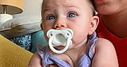 Paisley joined the competition — help win amazing prizes! adult, baby, blue_eyes, casual_clothing, child, closeup, comfort, couch, cushion, face, glasses, hand, holding, indoor, lamp, pacifier, parent, person, purple_dress, skin