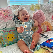 Paisley joined the competition — help win amazing prizes! baby, child, smiling, laughing, bow, plush_toy, book, quilt, couch, colorful, indoor, window, sunshine, happy, infant, clothing, daisy_pattern, person, portrait, playful