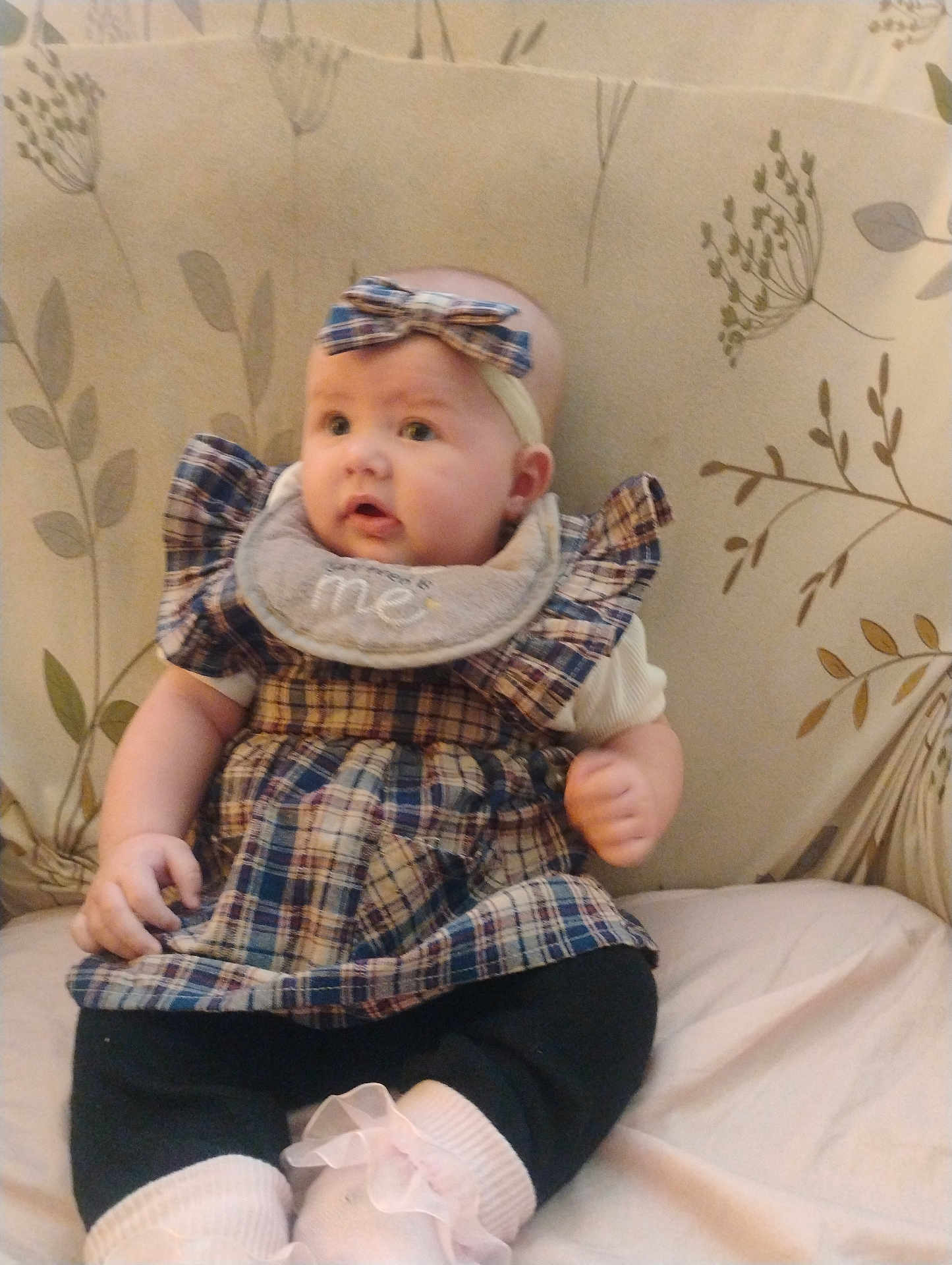 Luna is registered to the contest to win money with this photo: baby, child, plaid_dress, headband, bib, sitting, couch, floral_pattern, curious, indoors, soft_lighting, cute, infant, clothing, footwear, pink_socks, portrait, person, young_child, comfortable
