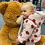 candid_photo, carpet_floor, child, cute, hooded_jacket, hugging, indoor, pacifier, pink_coat, plush, polka_dots, price_tag, retail_display, shelf, shoes, shopping, stuffed_animal, teddy_bear, toddler, toy_store