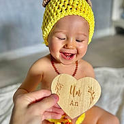 Lyam a rejoint le concours — aidez-le/la à gagner de superbes lots ! baby, blurred_background, child, closeup, crocheted_hat, cute, floor, hand, happy, indoors, infant, necklace, plaque, portrait, sitting, skin, smiling, warm_light, wooden_heart, yellow_hat
