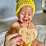 baby, blurred_background, child, closeup, crocheted_hat, cute, floor, hand, happy, indoors, infant, necklace, plaque, portrait, sitting, skin, smiling, warm_light, wooden_heart, yellow_hat