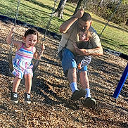 Harlee is registered to the contest to win money with this photo: child, toddler, man, swing, playground, outdoor, grass, trees, woodchips, sunlight, striped_dress, shorts, socks, shoes, tattoo, family, playing, summer, happy, fun