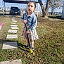 baseball_bat, car, child, curious_expression, daytime, denim_jacket, dress, footwear, grass, nature, outdoor, parking_area, path, person, shoes, sky, stepping_stones, sunlight, tree, yard