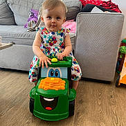 Luce a rejoint le concours — aidez-le/la à gagner de superbes lots ! baby, child, colorful, couch, cute, dress, floor, floral, furniture, green, happy, indoor, living_room, person, play, smiling_toy, toddler, toy, tractor, wooden_floor