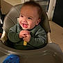 baby, child, high_chair, smile, happy, indoor, toy, green_sweater, hands, mouth_open, face, chair, plastic_tray, kitchen, door, floor_tile, evening, snack, playtime, cute