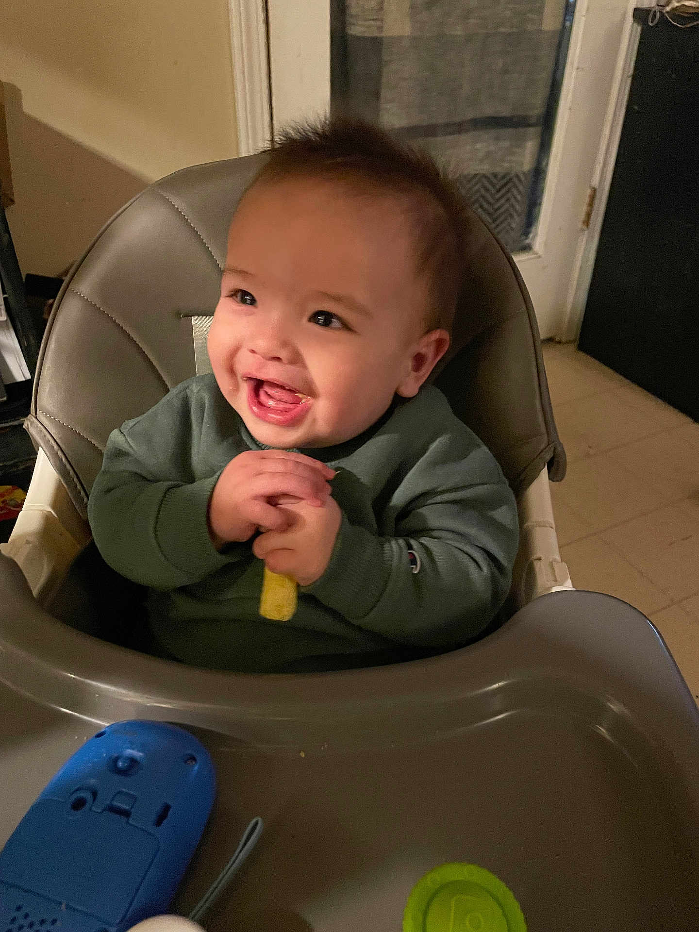 Riot is registered to the contest to win money with this photo: baby, child, high_chair, smile, happy, indoor, toy, green_sweater, hands, mouth_open, face, chair, plastic_tray, kitchen, door, floor_tile, evening, snack, playtime, cute