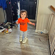 Mark is registered to the contest to win money with this photo: toddler, child, orange_shirt, oversized_shoes, sneakers, indoor, flooring, door, shoe_rack, shoes, clothing, person, standing, expression, light_switch, baby_gate, wall, plastic_bottle, home, curious