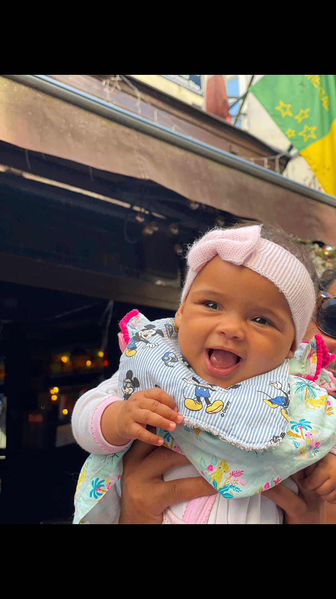 Mountabih Garrami joined the competition — help win amazing prizes! baby, child, smiling, headband, bib, mickey_mouse, hand, person, outdoor, building, flag, clothing, happy, portrait, cute, infant, holding, face, colorful, joyful
