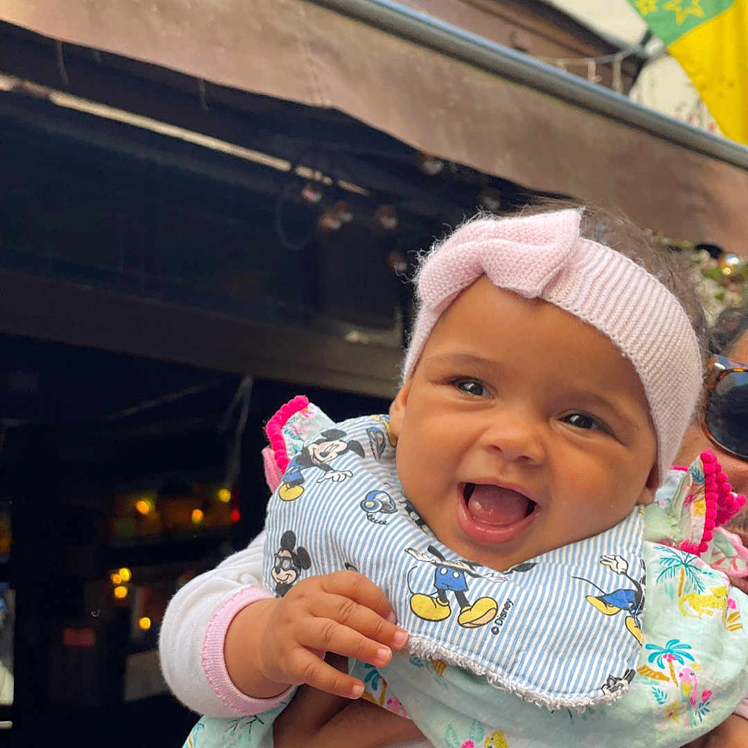 Lina Garrami a rejoint le concours — aidez-le/la à gagner de superbes lots ! apparel, baby, background, bib, child, clothing, colorful, cute, face, flag, hand, happy, headband, holding, infant, mickey_mouse, outdoor, person, portrait, smiling