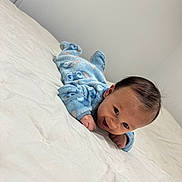Jaden is registered to the contest to win money with this photo: baby, blue_onesie, smiling, lying_down, bed, white_bedspread, infant, happy, tummy_time, indoors, child, cute, face, person, cozy, minimalist, soft_lighting, newborn, young_child, sleepwear