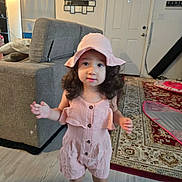 Eliana joined the competition — help win amazing prizes! child, toddler, girl, pink_hat, pink_outfit, romper, socks, curly_hair, standing, portrait, smiling, indoors, living_room, sofa, rug, door, home, furniture, fashion, photography