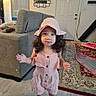 child, toddler, girl, pink_hat, pink_outfit, romper, socks, curly_hair, standing, portrait, smiling, indoors, living_room, sofa, rug, door, home, furniture, fashion, photography