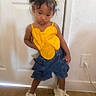 blue_shorts, casual_clothing, child, child_pose, cute_hairstyle, door, footwear, hair_clips, hair_puffs, home_interior, indoor, pensive_expression, posing, power_outlet, standing, white_sneakers, white_wall, wooden_floor, yellow_top, young_girl