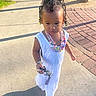 beaded_necklaces, bracelet, child, concrete, curly_hair, daytime, green_grass, hair_accessory, keys, outdoor, park, person, playful, shadow, sidewalk, sunlight, toddler, tree, walking, white_dress