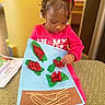 artwork, braids, child, colorful, craft, cushion, cute, decor, feet, focused, hair_accessories, hand, indoor, paper, red_clothing, sitting, table, tissue_paper, toddler, yarn