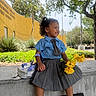 Aviora is registered to the contest to win money with this photo: toddler, child, smiling, yellow_flowers, blue_shirt, gray_skirt, curly_hair, backpack, concrete_ledge, outdoor, greenery, tree, building, sunny, side_profile, happy, casual_clothing, sneakers, daytime, urban