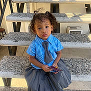 Aviora is registered to the contest to win money with this photo: toddler, child, curly_hair, pigtails, blue_shirt, gray_tie, gray_skirt, concrete_stairs, outdoor, holding_object, curious_expression, casual, portrait, daylight, person, young_child, sitting, clothing, fashion, cute