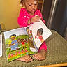 barefoot, book, child, colorful, cozy, curly_hair, expression, foot, hair_beads, illustration, indoors, pajamas, patterned, pink_clothing, reading, sitting, storybook, table, toddler, warm_lighting