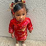 child, toddler, portrait, standing, red_outfit, floral_pattern, traditional_clothing, headpiece, hair_ornament, jewelry, face, serious_expression, indoor, textured_wall, tile_floor, shoes, fashion, cultural_attire, full_body, photography