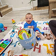 Henderson joined the competition — help win amazing prizes! baby, child, playmat, toys, basket, blue_onesie, indoor, floor, plant, furniture, carpet, stairs, animal_print, smiling, happy, person, colorful, toy_car, reading, playtime