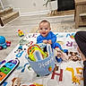 baby, child, playmat, toys, basket, blue_onesie, indoor, floor, plant, furniture, carpet, stairs, animal_print, smiling, happy, person, colorful, toy_car, reading, playtime