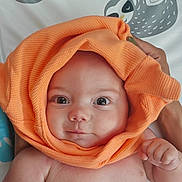 Luca is registered to the contest to win money with this photo: baby, infant, orange_cloth, face, eyes, hand, skin, expression, cute, adorable, hood, fabric, texture, sloth_illustration, background, bed, person, portrait, newborn, child