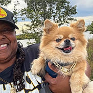 Reeses is registered to the contest to win money with this photo: accessory, black_clothing, cloud, dog, fur, gold_chain, grass, hand, happy, hat, lake, nature, outdoor, person, pet, pomeranian, portrait, sky, smile, tree