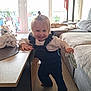 toddler, child, smiling, standing, indoor, living_room, sofa, cat, blanket, window, natural_light, sock, barefoot, hair_tie, denim_overalls, white_shirt, floor, table, playful, happy