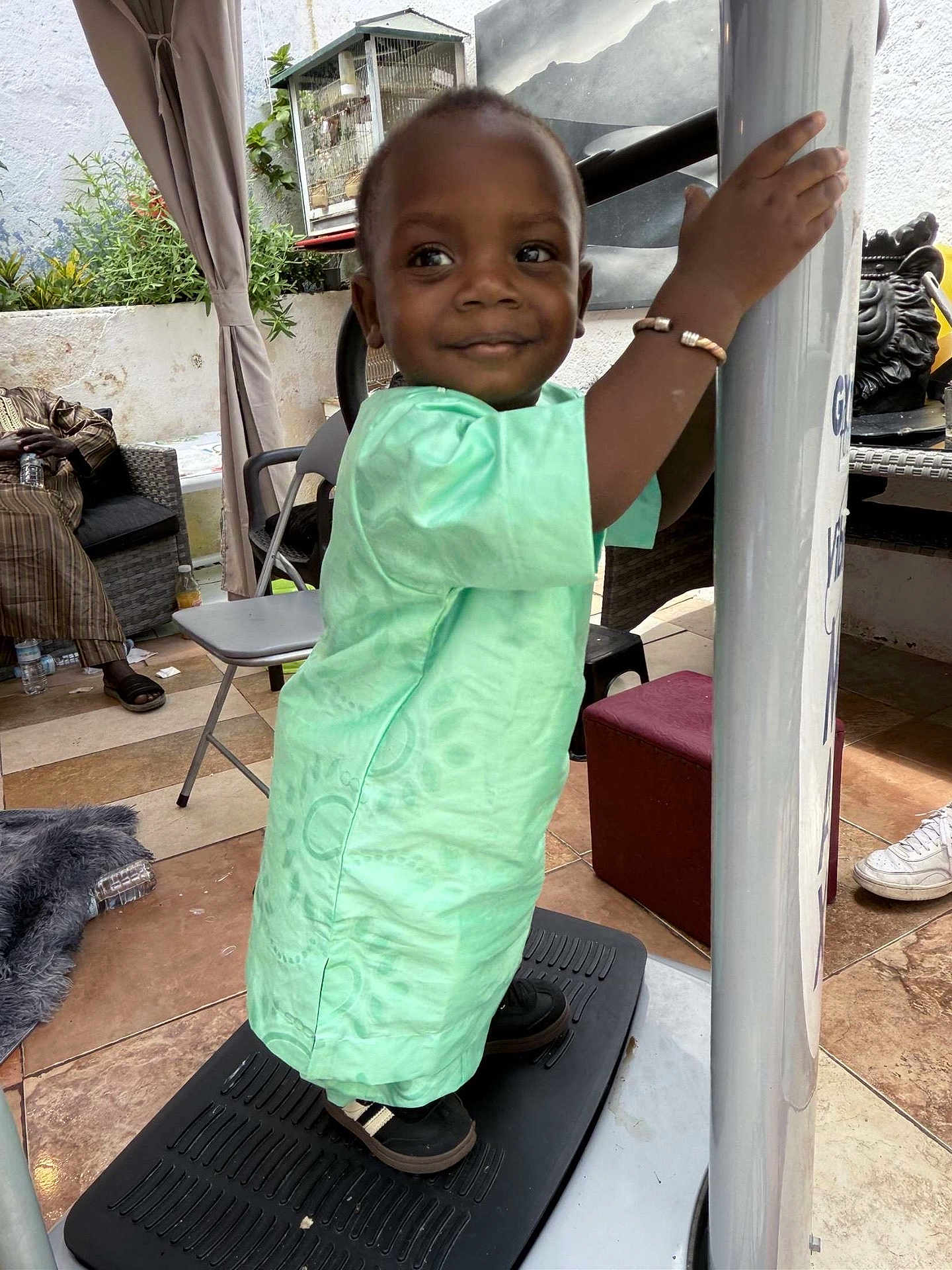 Malick a rejoint le concours — aidez-le/la à gagner de superbes lots ! child, toddler, smile, green_clothing, bracelet, platform, pole, outdoor, plants, chair, person, shoe, floor_tile, curtain, cage, adult, casual, happy, portrait, home
