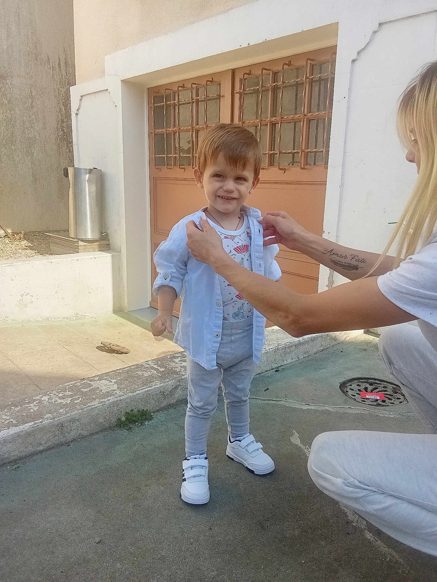 Teo participe au concours pour gagner de l'argent avec cette photo : child, boy, smiling, shirt, pants, sneakers, outdoor, concrete, door, window, adult, tattoo, light, daylight, casual_clothing, person, standing, helping, happy, curly_hair