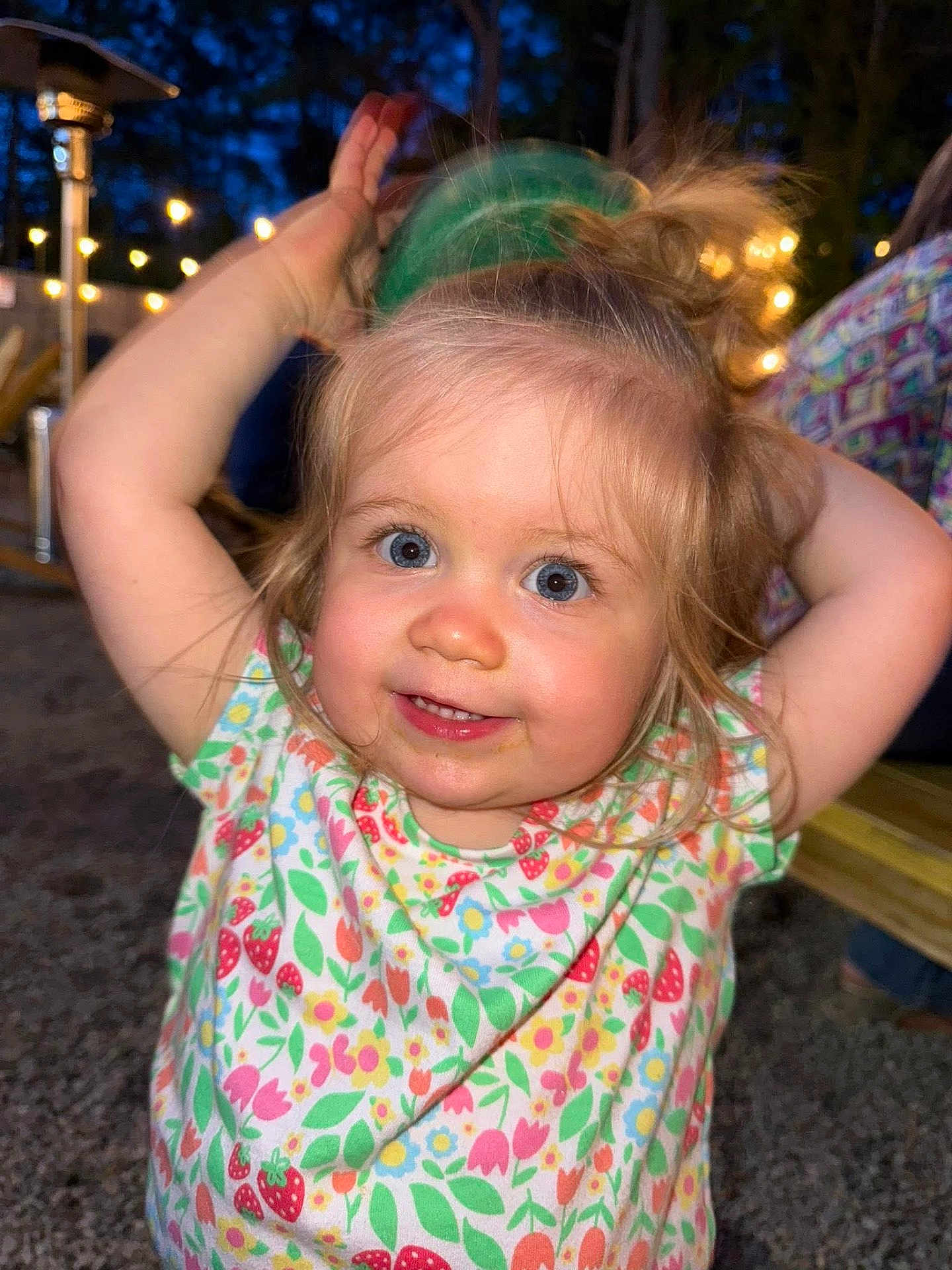 Lily is registered to the contest to win money with this photo: child, toddler, smiling, blue_eyes, blonde_hair, floral_shirt, outdoor, night, string_lights, playing, happy, cute, person, portrait, face, arm_raised, green_cup, hair_up, background_blur, fun
