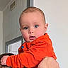 adult_hand, baby, bright_eyes, child, closeup, curious, cute, expression, face, hand, head, holding, indoor, infant, orange_clothing, person, portrait, skin, warm_clothing, young_child