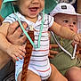 baby, infant, child, hat, bucket_hat, smiling, curious, striped_onesie, green_onesie, adult_hand, toy, sitting, patterned_clothing, cute, indoor, person, footwear, sock, happy, holding