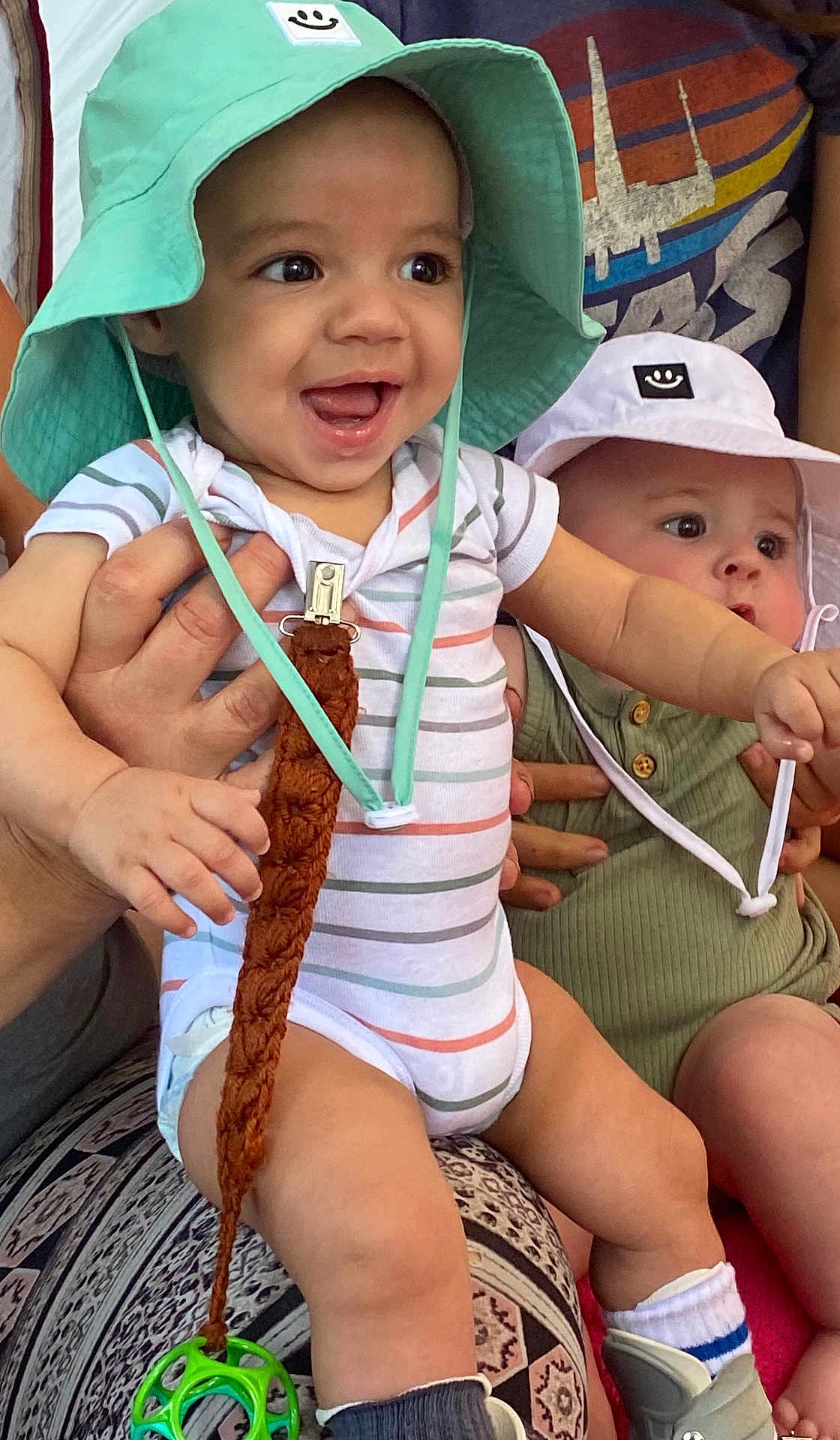 Valerian is registered to the contest to win money with this photo: baby, infant, child, hat, bucket_hat, smiling, curious, striped_onesie, green_onesie, adult_hand, toy, sitting, patterned_clothing, cute, indoor, person, footwear, sock, happy, holding