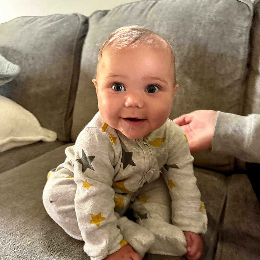 Leah is registered to the contest to win money with this photo: baby, child, onesie, stars, couch, gray, hand, indoor, smiling, happy, infant, cute, person, seated, plush, portrait, cozy, young, face, home