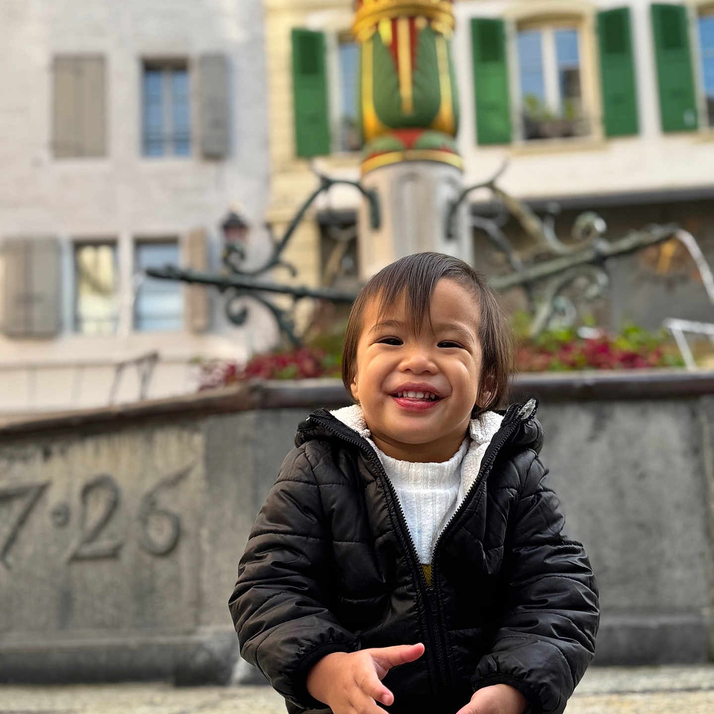 Toahei a rejoint le concours — aidez-le/la à gagner de superbes lots ! beige_pants, black_jacket, building, candy, casual_clothing, child, cute, daylight, fountain, green_shutters, happy, outdoor, person, playful, portrait, smile, stone_pavement, toddler, urban, window