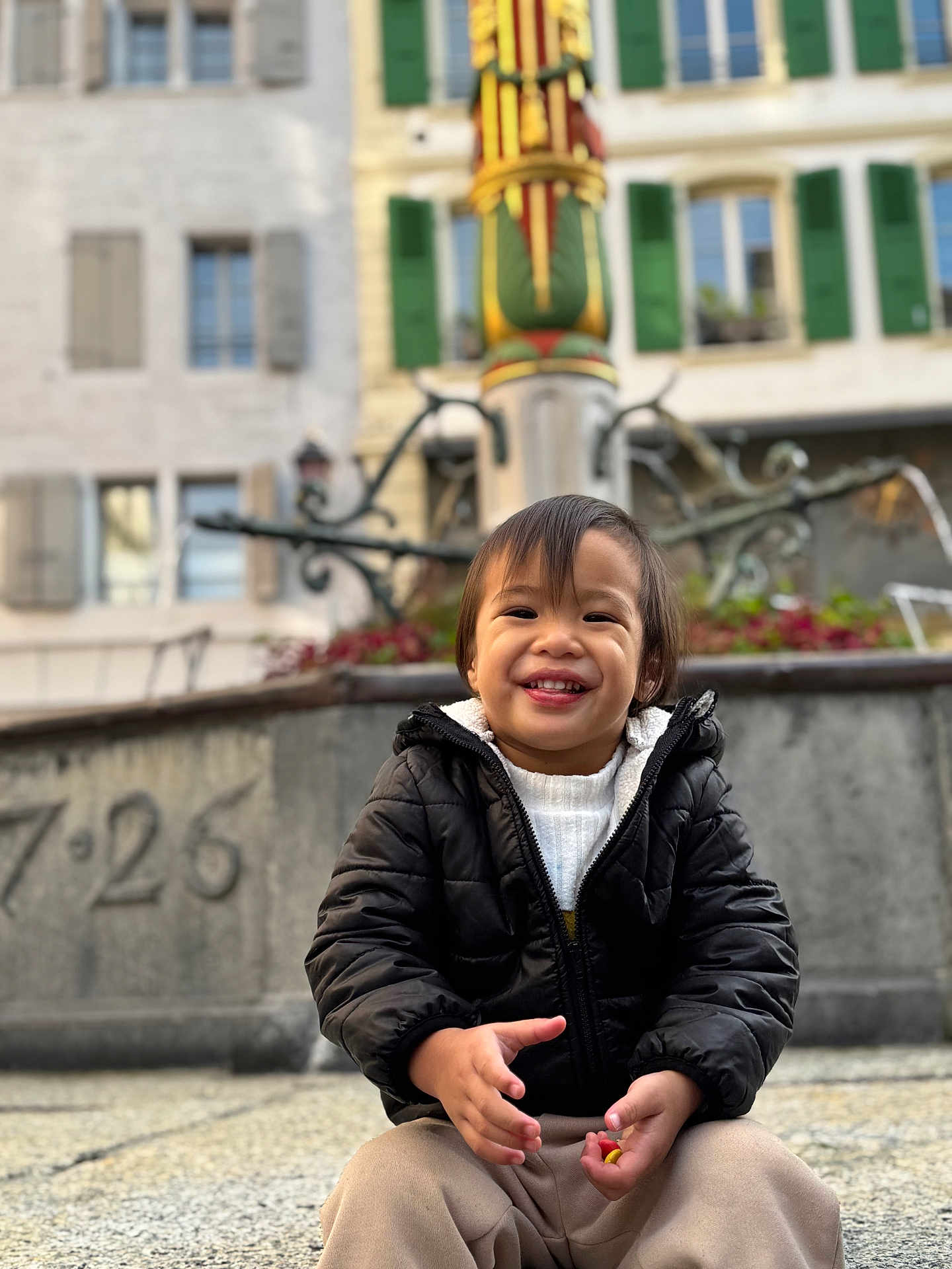 Toahei a rejoint le concours — aidez-le/la à gagner de superbes lots ! toddler, child, smile, black_jacket, beige_pants, candy, fountain, stone_pavement, outdoor, building, window, green_shutters, happy, person, casual_clothing, daylight, urban, playful, cute, portrait