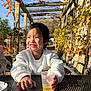 child, smiling, white_sweater, plastic_cup, outdoor, autumn, vines, wooden_pergola, sunlight, metal_table, happy, portrait, young_child, nature, daylight, casual_clothing, seated, table, plant, relaxed