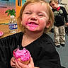 child, toddler, cupcake, pink_frosting, smiling, messy, classroom, water_bottle, table, black_shirt, happy, indoor, person, young_child, playful, food, celebration, fun, background, curious