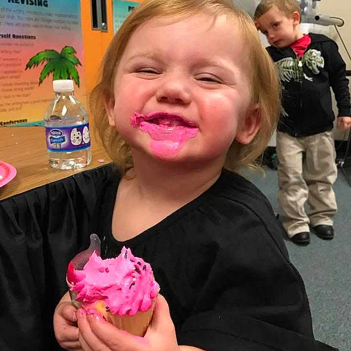 Zoey is registered to the contest to win money with this photo: child, toddler, cupcake, pink_frosting, smiling, messy, classroom, water_bottle, table, black_shirt, happy, indoor, person, young_child, playful, food, celebration, fun, background, curious