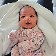 Shiloh Rose is registered to the contest to win money with this photo: baby, infant, blanket, onesie, floral_pattern, person, face, eyes, hand, lap, clothing, black_pants, wooden_furniture, indoor, cute, soft_texture, warm, newborn, portrait, child