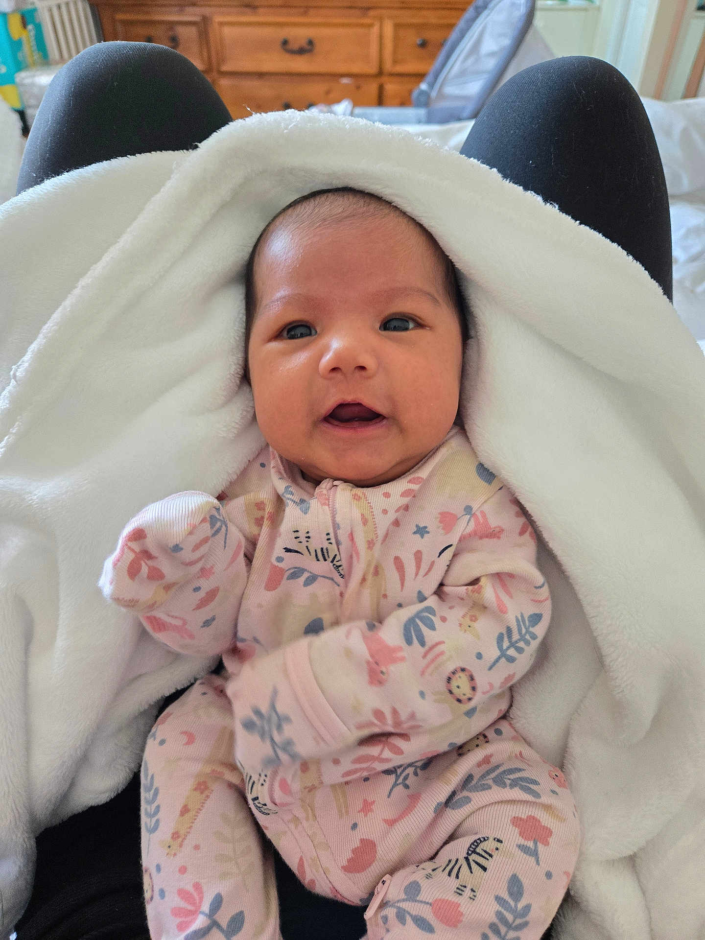 Shiloh Rose is registered to the contest to win money with this photo: baby, infant, blanket, onesie, floral_pattern, person, face, eyes, hand, lap, clothing, black_pants, wooden_furniture, indoor, cute, soft_texture, warm, newborn, portrait, child