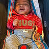 baby, child, infant, car_seat, red_hoodie, striped_clothing, gray_pants, blanket, seatbelt, smiling, happy, indoor, person, cute, portrait, clothing, toddler, comfort, holding, strap