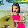 child, girl, braided_hair, pink_dress, playground, dinosaur_ride, outdoor, daylight, park, play_equipment, smiling, happy, person, fence, sidewalk, grass, trees, sunlight, holding, recreation
