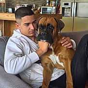 Taïro participe au concours pour gagner de l'argent avec cette photo : young_man, dog, boxer_dog, couch, white_hoodie, indoor, kitchen, refrigerator, microwave, wooden_table, gray_furniture, casual, pet, friendship, relaxation, portrait, home_interior, seated, comfort, companion