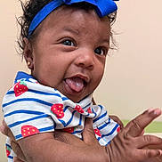 Khalani joined the competition — help win amazing prizes! baby, child, face, tongue, headband, blue_bow, curly_hair, earrings, striped_shirt, strawberry_pattern, hand, holding, smile, playful, indoor, person, cute, portrait, infant, skin